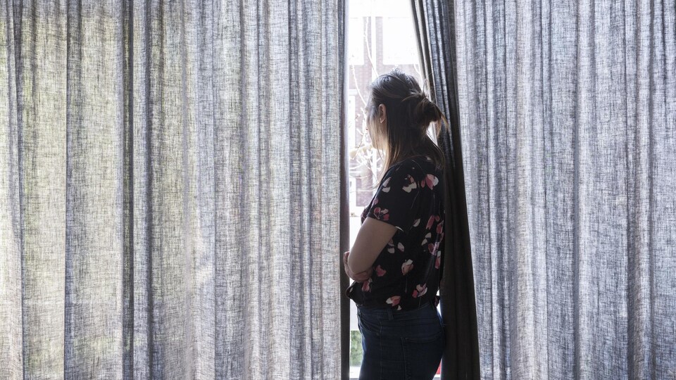 Une femme regarde l'extérieur par l'ouverture des rideaux, debout devant une fenêtre.
