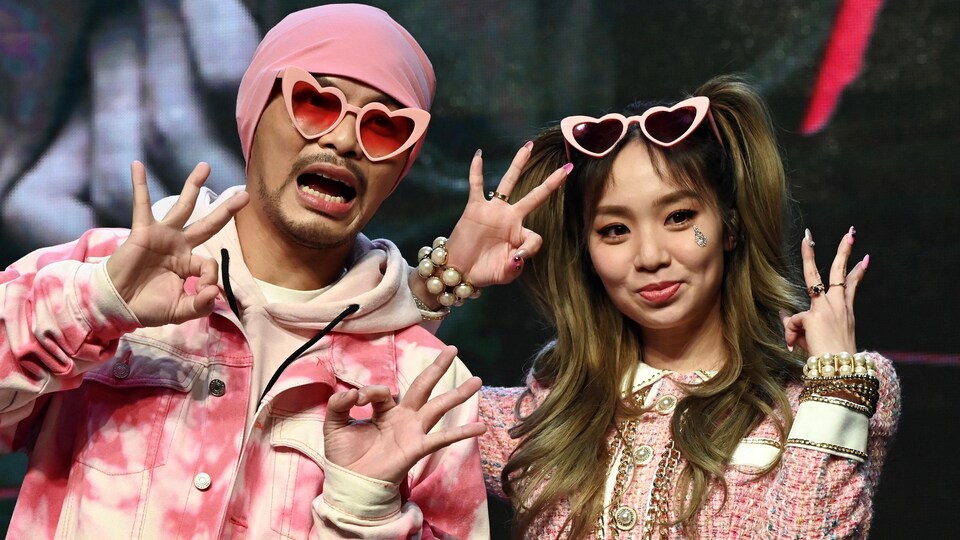 Les deux chanteurs portent des vêtements roses et des lunettes roses. Leurs mains forment le signe «OK», en formant un cercle avec le pouce et l'index, et en tenant les autres doigts droits loin de la paume.