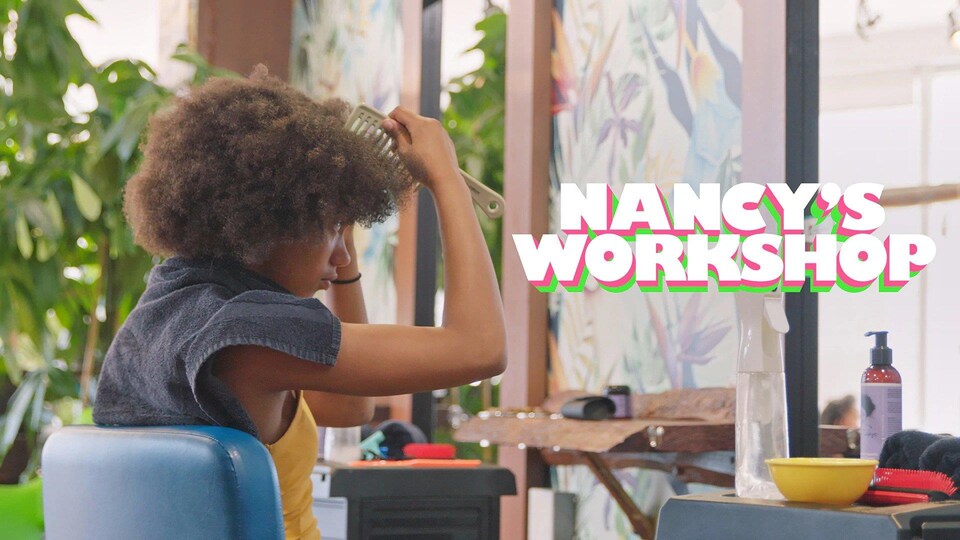 Image promotionnelle dans laquelle on voit une jeune femme travailler ses cheveux frisés serrés dans au miroir d'un salon de coiffure