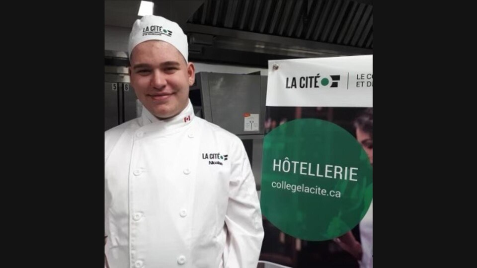 Nicolas Ouellet porte sa tenue de chef cuisinier et sa toque dans une cuisine à côté d'une affiche du Collège La Cité.