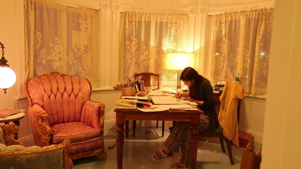 Une jeune femme dessine à un bureau, entourée de meubles antiques.