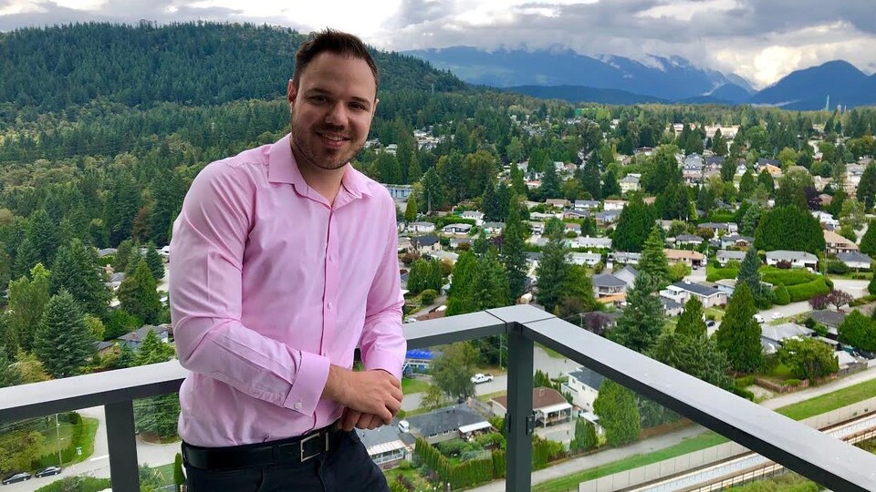 Un homme est accoté sur le garde-fou de sa terrasse, avec une vue sur la ville.