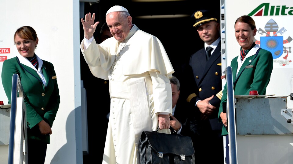 Pope Francis waves to people as he exits a plane upon arrival at Rome's Fiumicino Airport on September 19, 2015.  Two flight attendants surround him and a pilot behind him. 