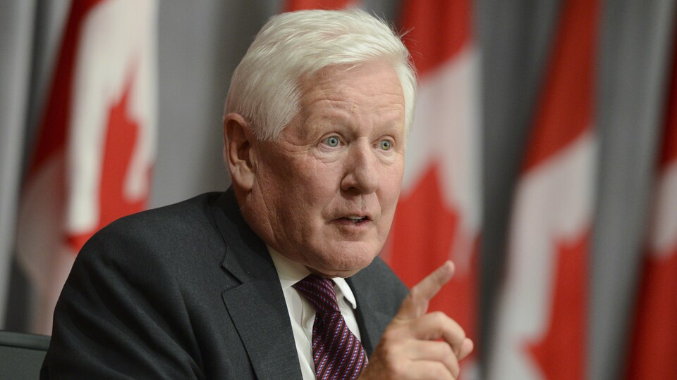 Bob Rae parle, assis devant des drapeaux canadiens.