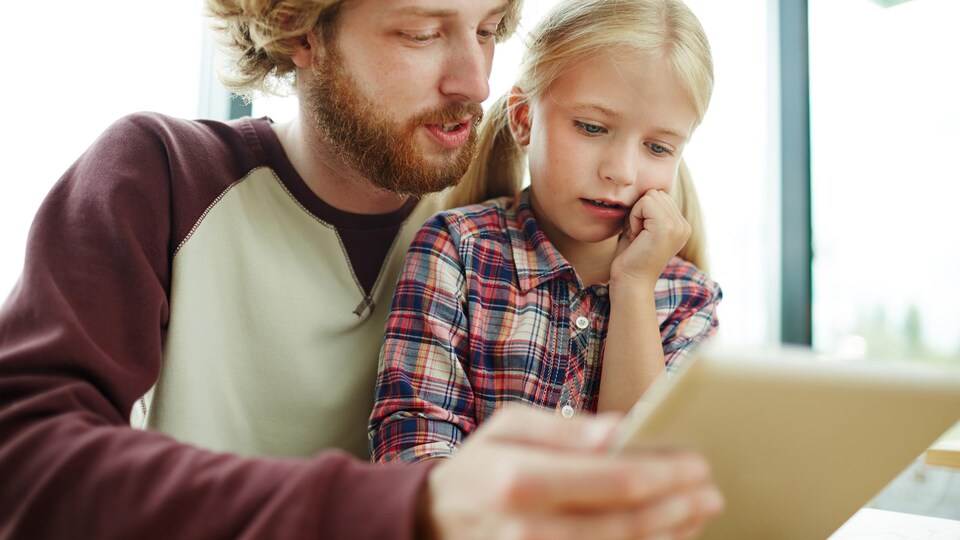 Un père tient une tablette électronique et la montre à sa fille.