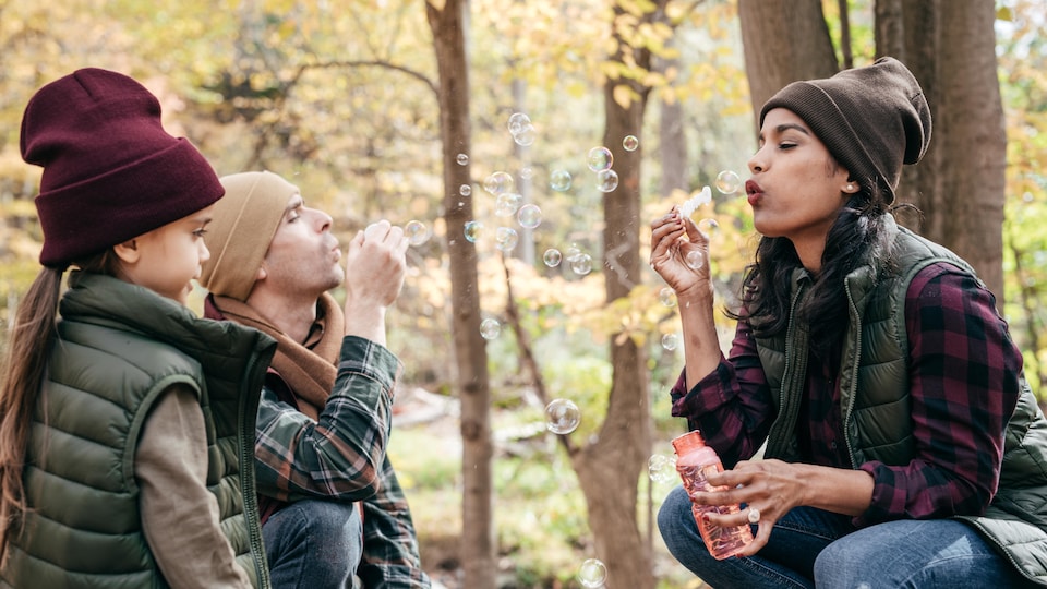 Une petite fille et ses parents, dans le bois, habillés pour l'automne, jouent à faire des bulles. 