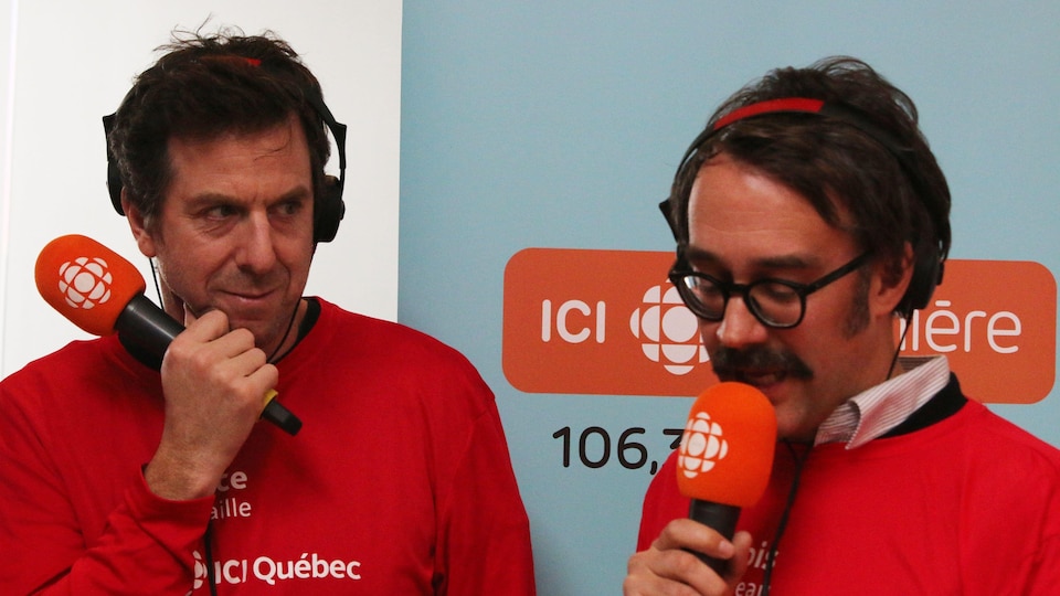 Deux hommes parlent au micro avec en arrière plan un affiche bleu pâle. Ils portent tout deux des chandails rouges.