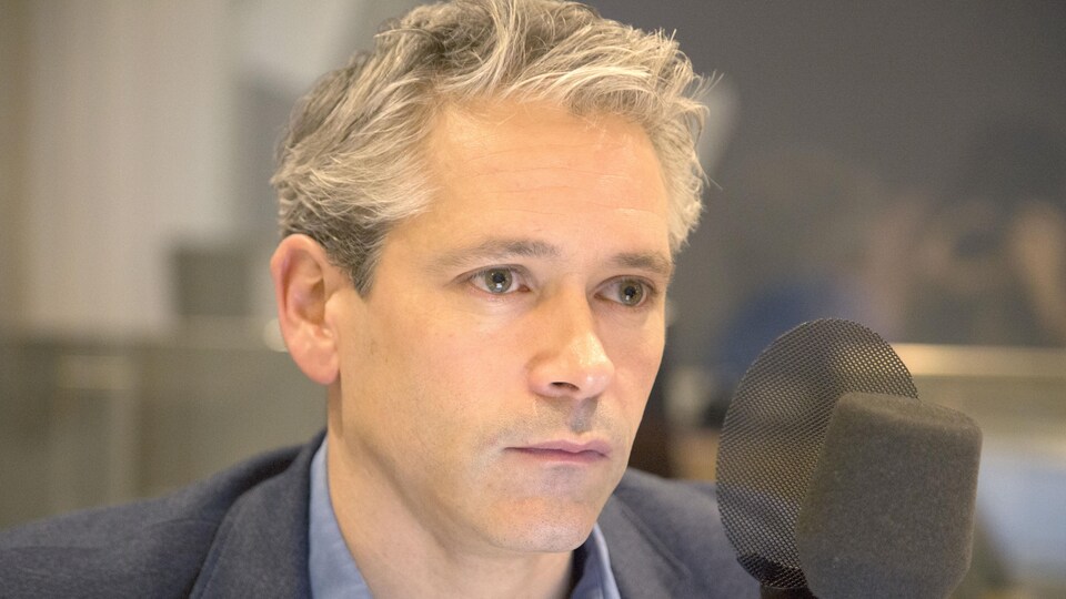 Il est assis devant un micro en regardant devant lui.