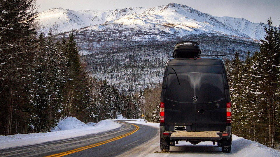 On voit un véhicule fourgonnette sur une route, immobile. Devant, il y a des montagnes enneigées. 