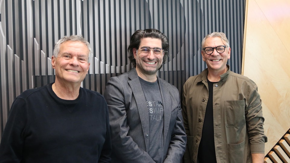 Trois hommes prennent la pose en souriant devant un logo de Radio-Canada dans un studio de radio. 