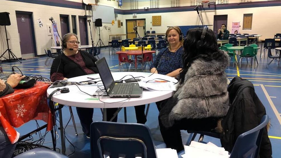 Deux invitées au micro d'une animatrice de radio à l'intérieur d'un gymnase scolaire.
