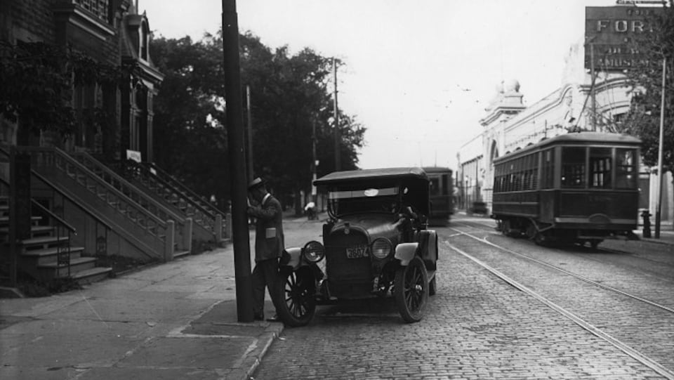 Photo en noir et blanc montrant une rue pavée sur laquelle on voit une automobile ancienne et un tramway.