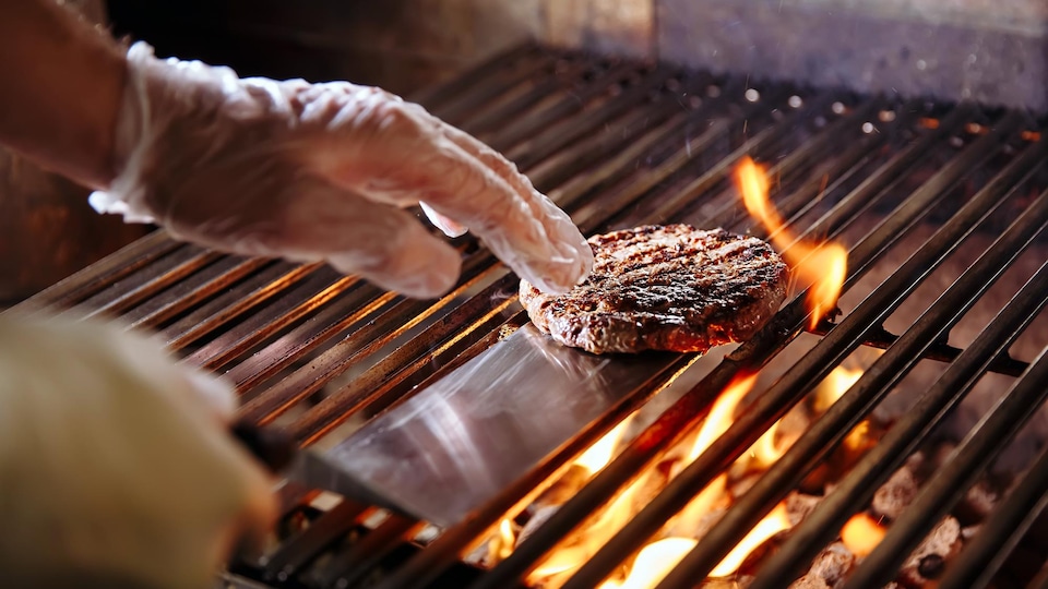 Munie d'une spatule métallique, une personne cuit une galette de viande hachée sur un barbecue.