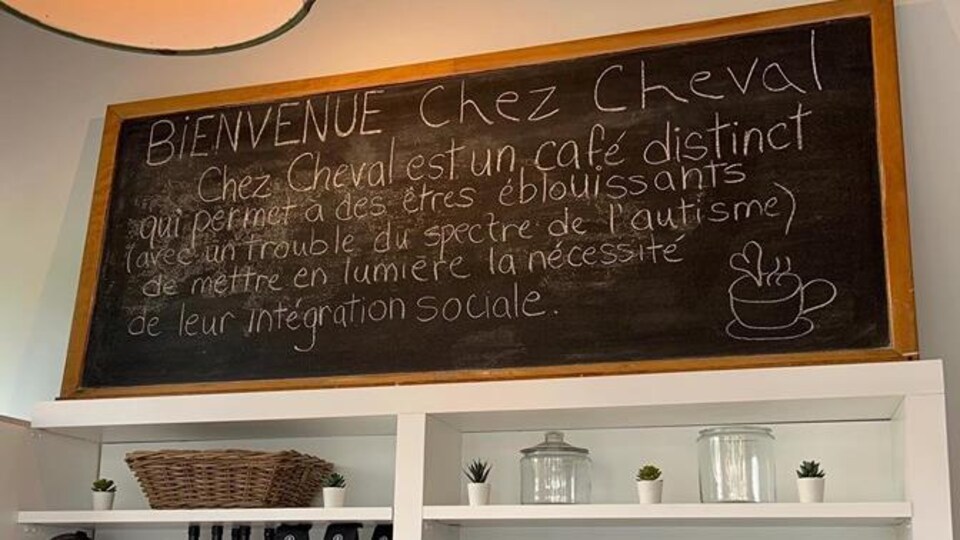 Photo d'un grand tableau posé sur des étagères sur lequel il est inscrit : « Bienvenue Chez Cheval. Chez Cheval est un café distinct qui permet à des êtres éblouissants (avec un trouble du spectre de l'autisme) de mettre en lumière la nécessité de leur intégration sociale. »
