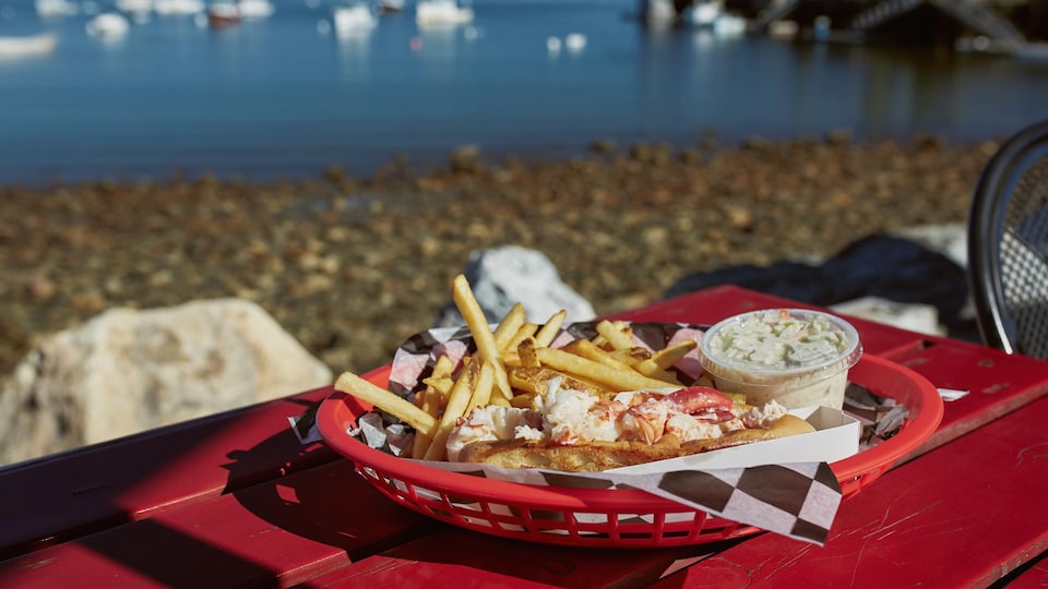 Un plat de frites, salade de choux et d'une guédille est posé sur une table rouge avec des bâteaux sur l'eau au loin.