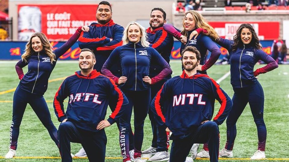 Des hommes et des femmes posent sur un terrain de football en tenue de cheerleaders des Alouettes de Montréal.