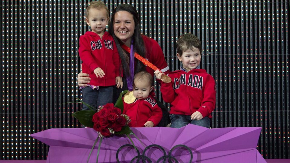 Christine Girard pose sur la plus haute marche d'un podium olympique avec ses trois petits garçons avec une médaille d'or et une médaille de bronze autour du cou.
