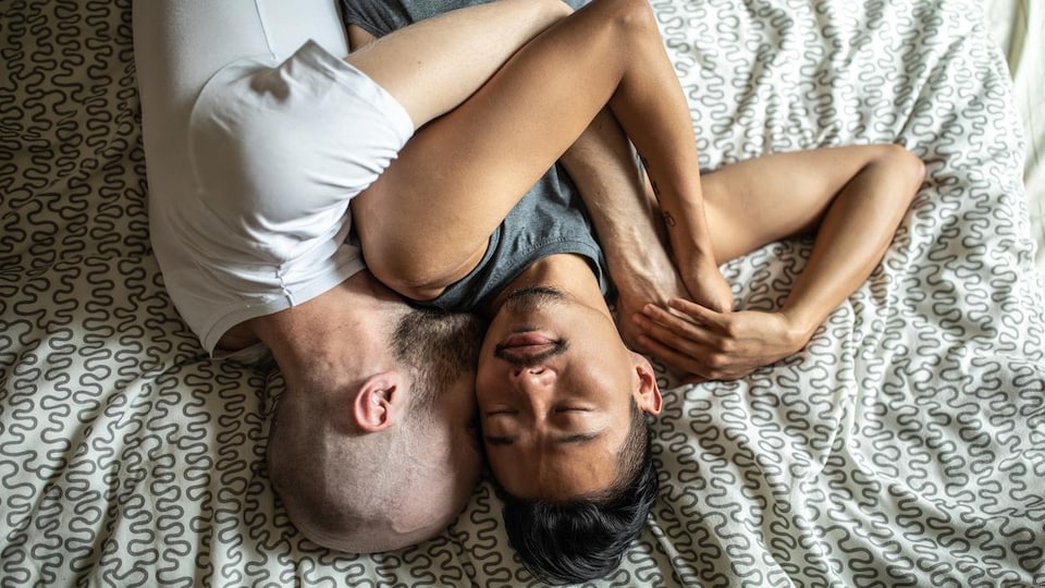 Deux hommes se prennent dans les bras, habillés, sur un lit.