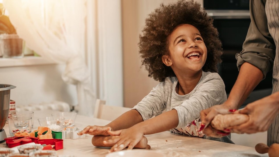 Une petite fille tient un rouleau à pâtisserie et fait un grand sourire à sa maman qui cuisine avec elle. 