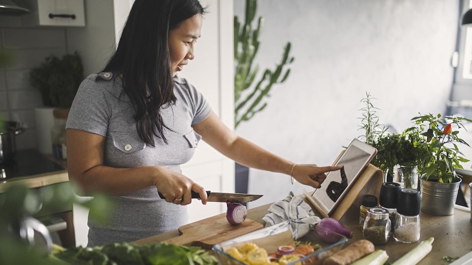Une femme coupe des légumes en regardant une recette sur une tablette.