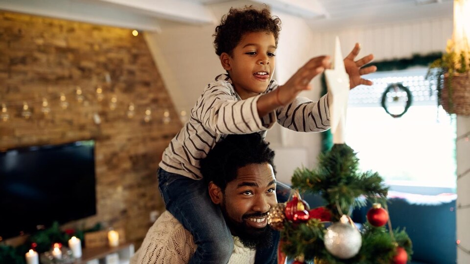 Un père tient son fils sur ses épaules qui installe l'étoile au sommet d'un sapin de Noël.