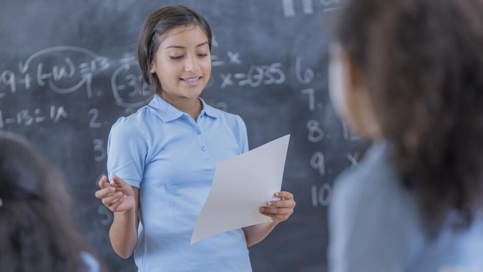 Une fille devant un tableau noir lit une feuille devant les autres élèves.