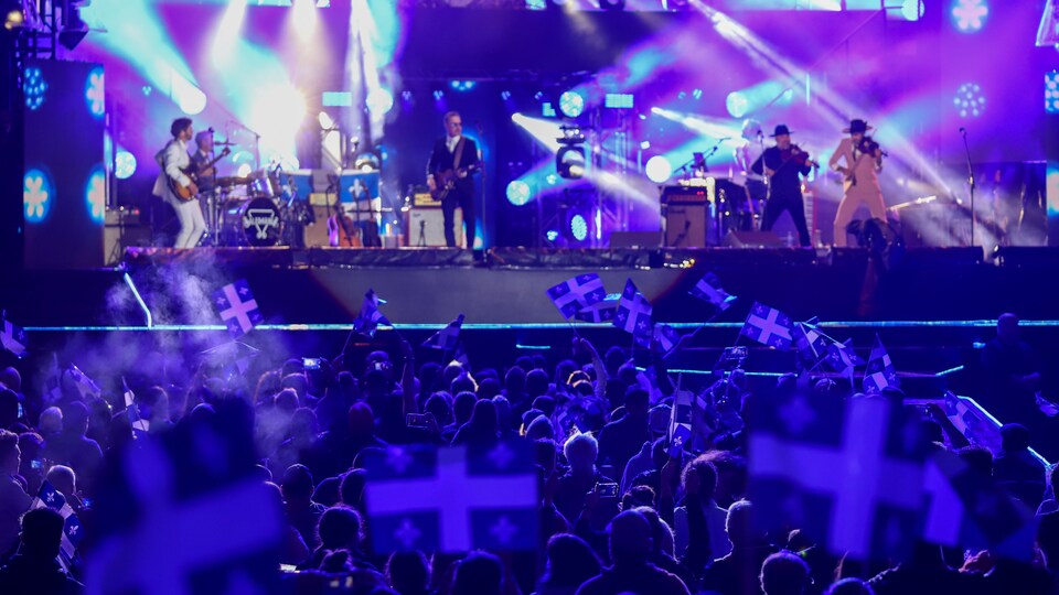 Un groupe joue sur scène lors du spectacle de la fête nationale Québec; plusieurs gens tiennent un drapeau du Québec dans la foule.