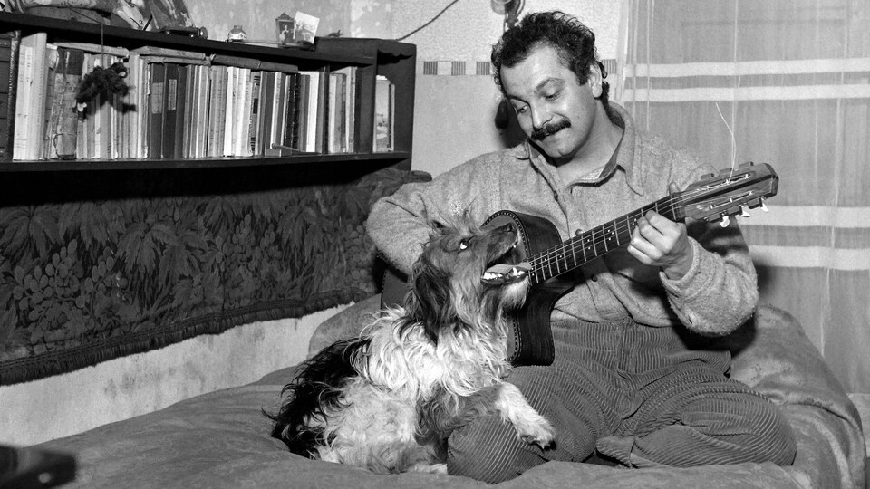 Il joue de la guitare, assis sur un lit avec un chien qui le regarde.