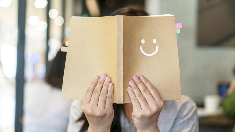 Une femme couvre son visage avec un livre ouvert devant elle sur la couverture duquel un bonhomme sourire est imprimé.