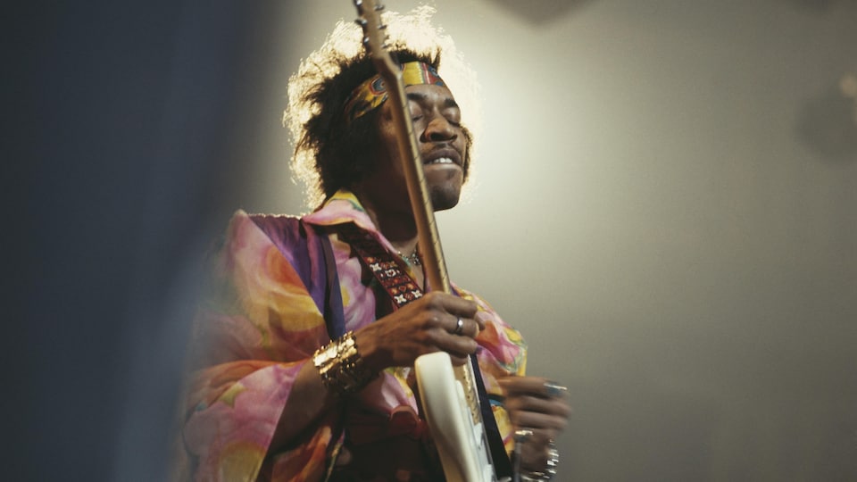 Jimi Hendrix sur scène, une guitare à la main, avec une chemise colorée et un bandeau dans les cheveux.