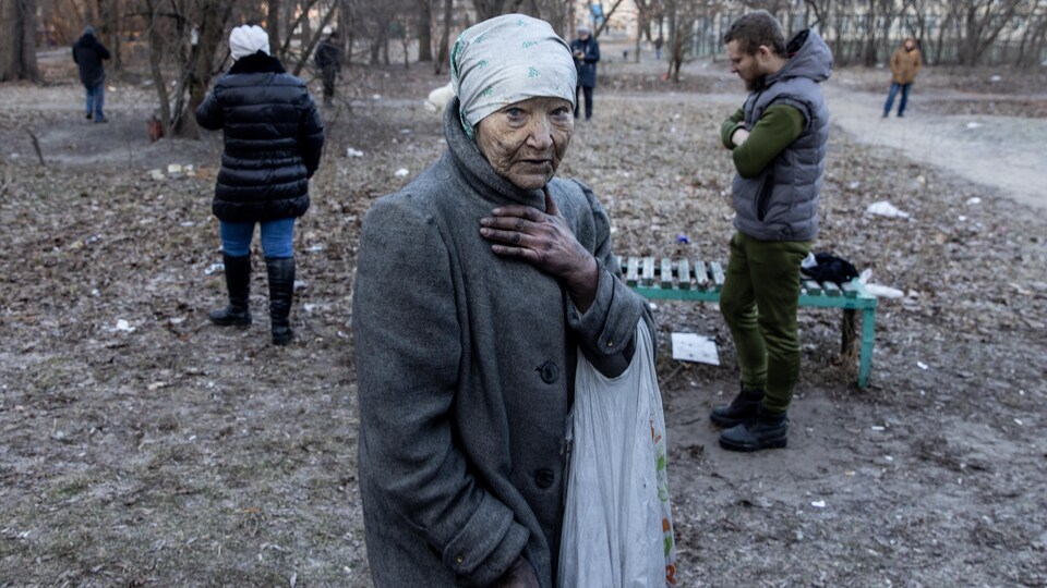 Une femme âgée se tient seule, les mains et le visage noircis, un sac plastique avec des affaires à son bras.