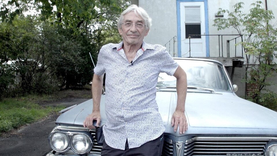 Il pose devant le capot d'une voiture Pontiac.