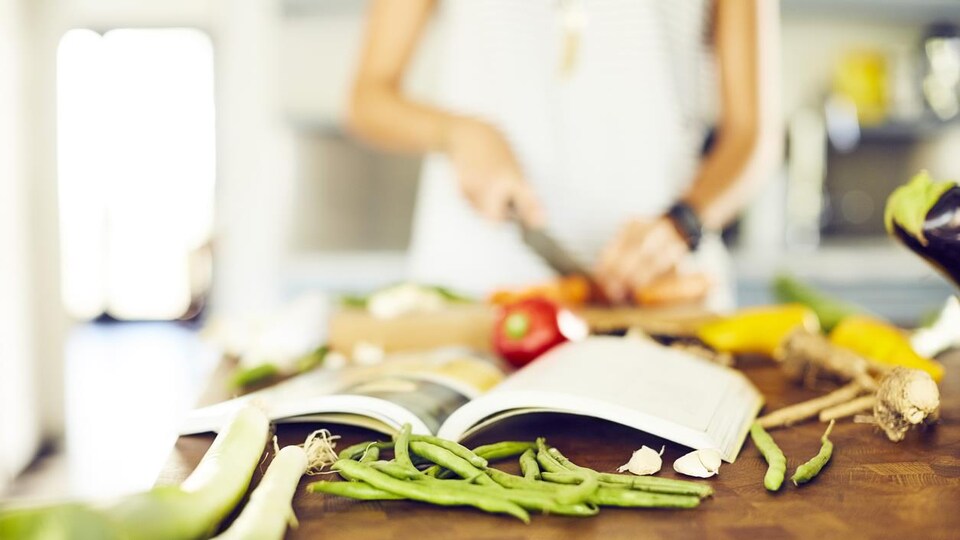 Une femme consulte un livre de recettes ouvert devant elle pendant qu'elle coupe des légumes. 