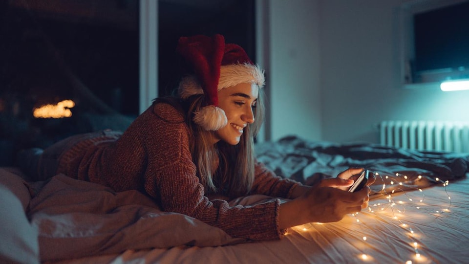 Une jeune femme allonger sur un lit avec un chapeau du père Noël et une guirlande lumineuse regarde son cellulaire. 