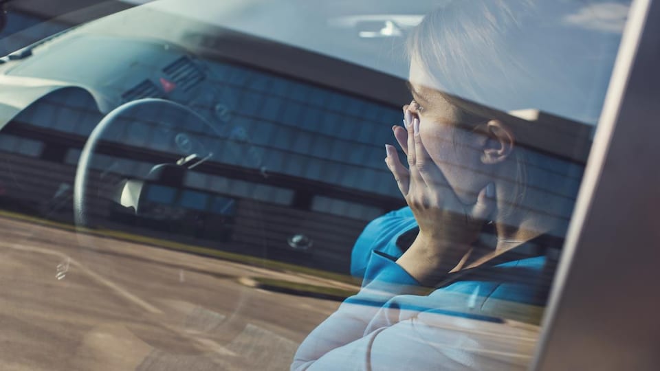 Une femme assise au volant de sa voiture se tient le visage dans les mains. 
