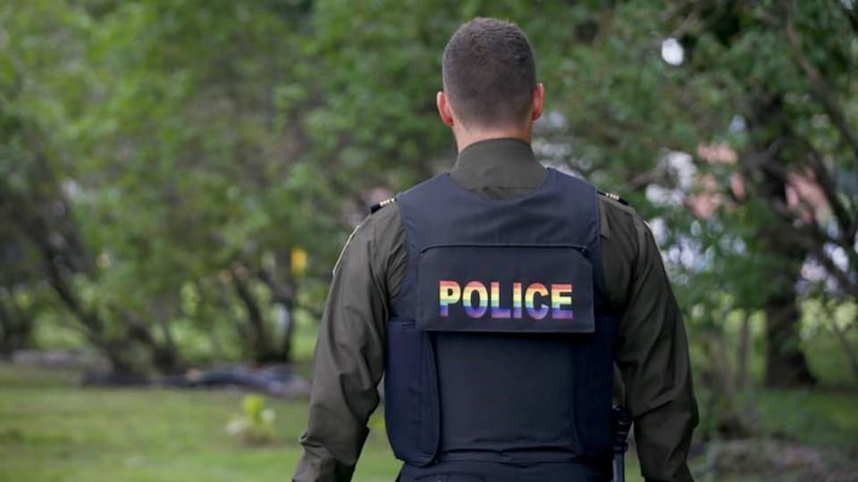 Un policier de dos porte un gilet pare-balles avec l'inscription "Police" inscrite aux couleurs du drapeau arc-en-ciel. 
