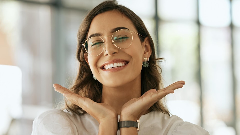 Une jeune femme avec des lunettes sourie et a placé ses mains sous son visage.