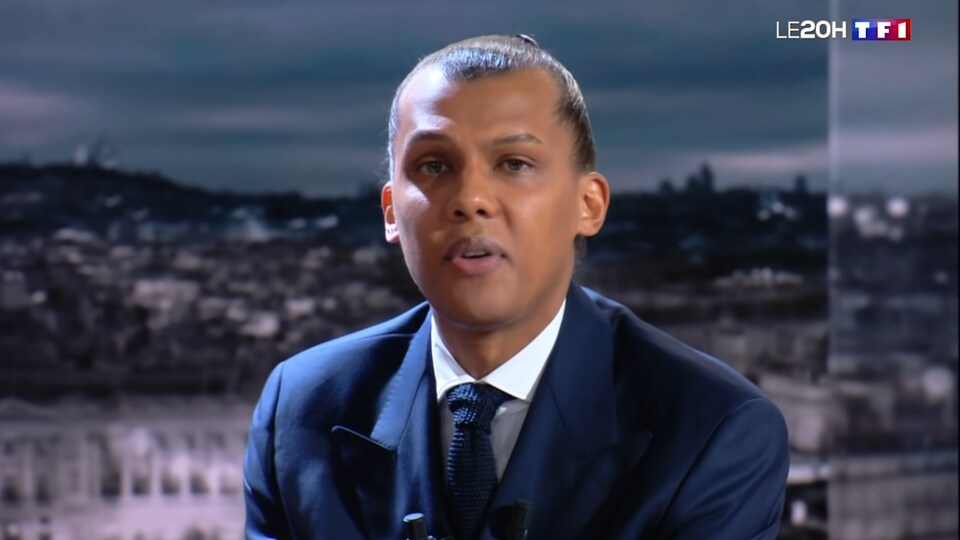 L'homme vêtu d'un complet chante alors qu'il est assis derrière une table sur un plateau de télévision.