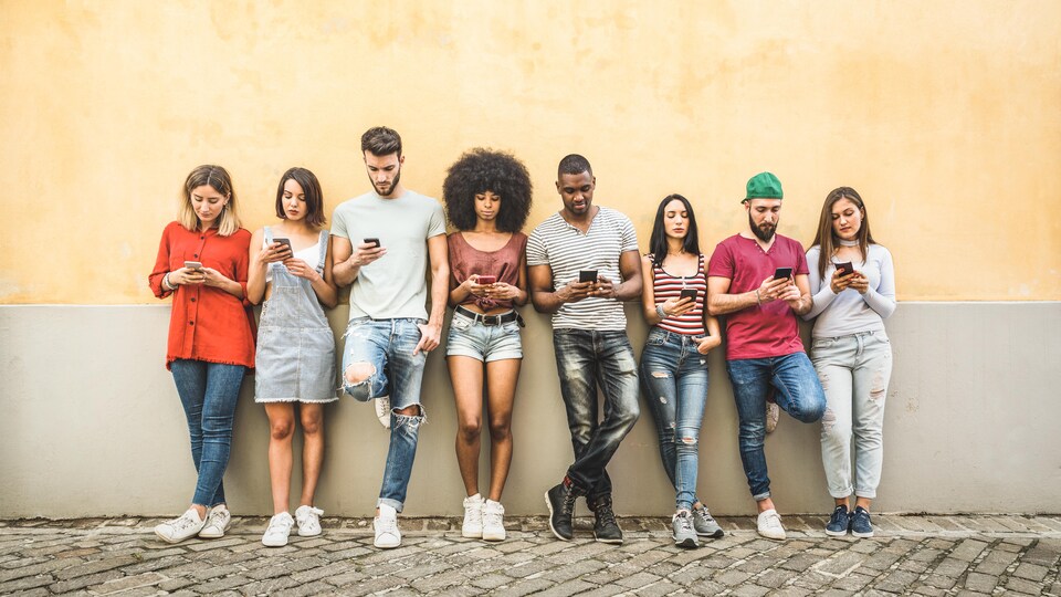 Un groupe de jeunes personnes accotées sur un mur, toutes sur leur téléphone cellulaire.