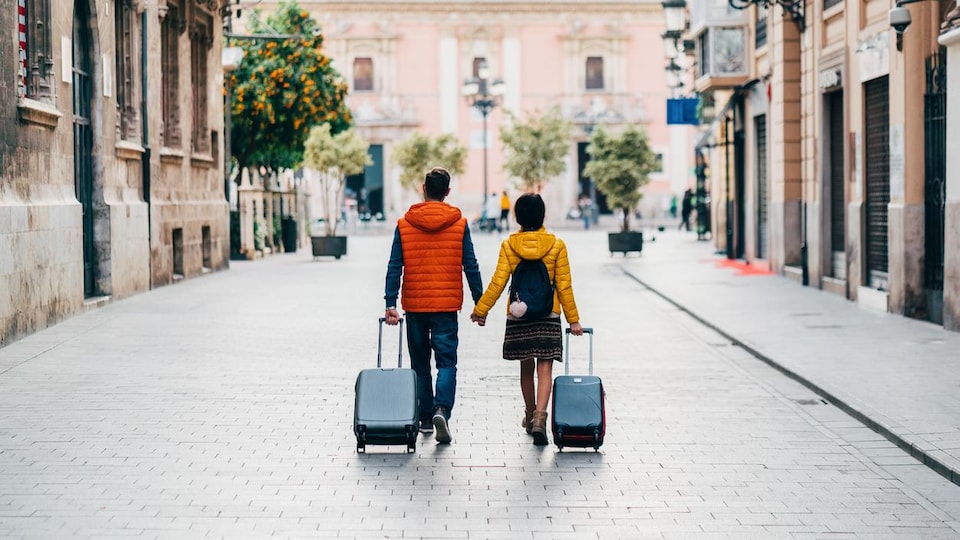 Un jeune couple marche en se tenant la main et en tirant ses valises dans les rues de Valence, en Espagne.