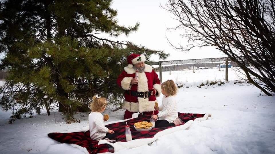 Dans un champ enneigé, le Père Noël fait un pique-nique. Il boit du lait et mange des biscuits avec deux fillettes.