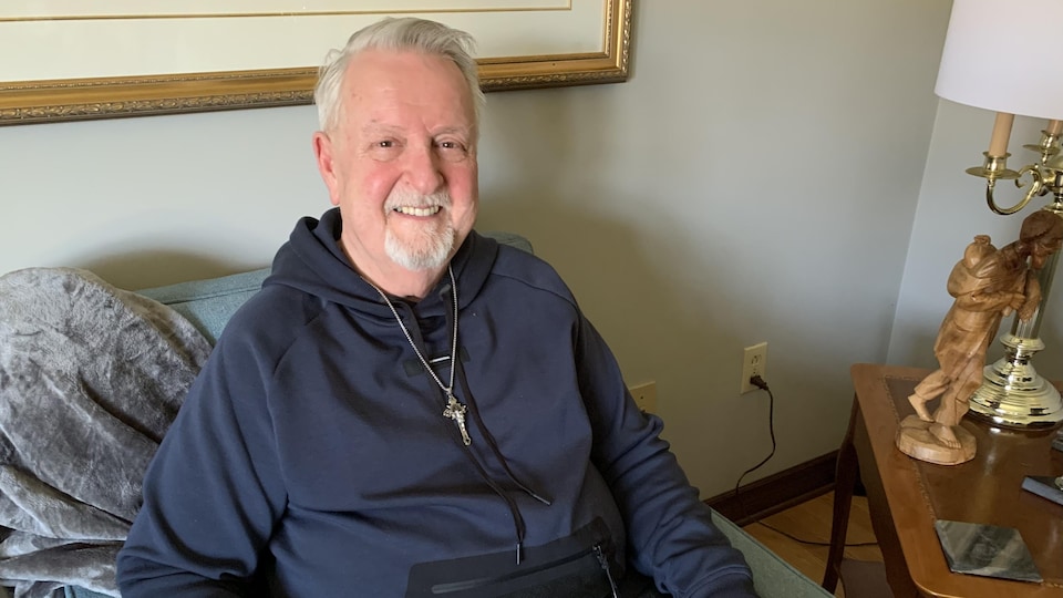 Père Robert Champagne du Sud-Ouest de l'Ontario, souriant, portant une chaîne avec une croix, est assis sur un fauteuil.
