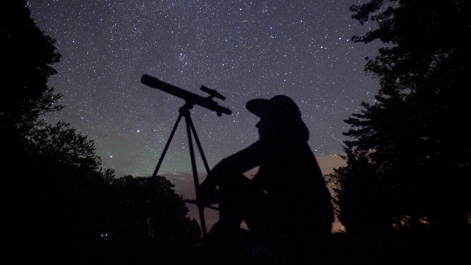 Une personne assise à côté de son télescope sous un ciel d'étoiles. 