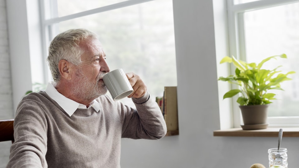 Un homme âgé en train de boire dans une tasse en regardant par la fenêtre.