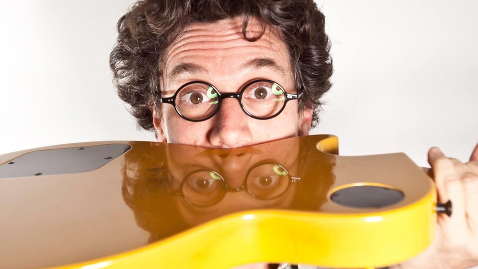 Le musicien à lunettes rondes est vu le visage appuyé sur l'envers d'une guitare jaune.