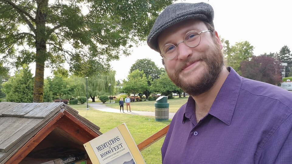 Un homme souriant tient un livre devant une petite bibliothèque dans un parc. 