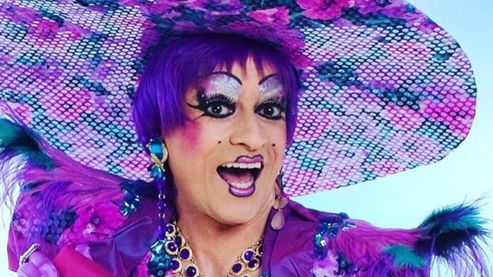 La drag queen Mado Lamotte est habillée en mauve avec un très grand chapeau fleuri.