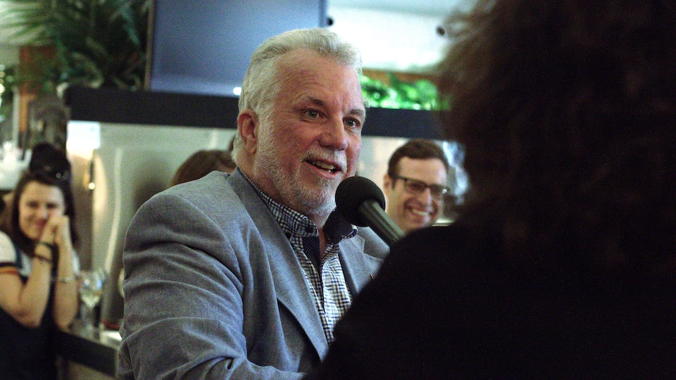 Philippe Couillard, derrière un micro noir, regarde Christiane Charette, de dos.