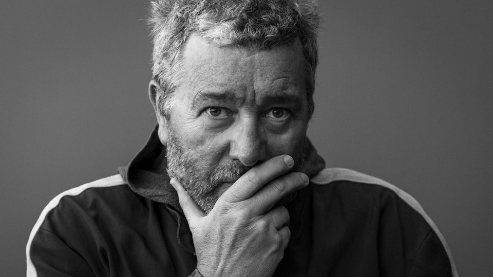 Portrait de Philippe Starck, barbe et cheveux courts, se tenant la bouche avec la main. La photo est en noir et blanc.