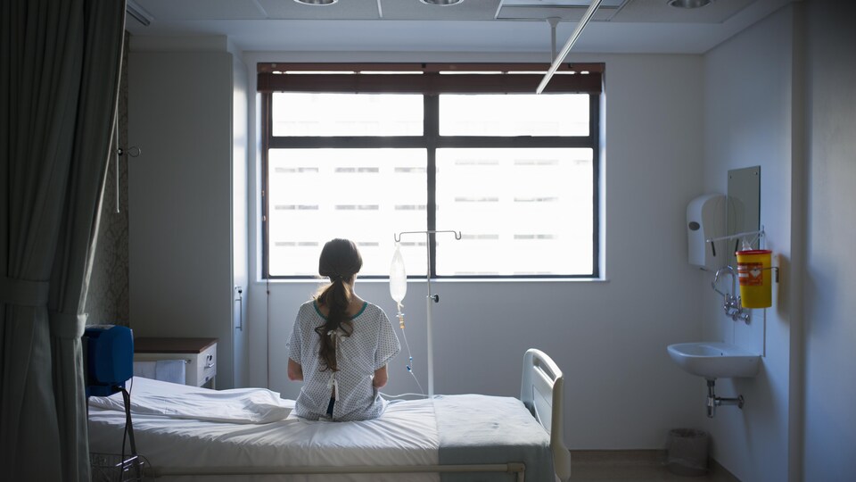 Une patiente de dos, assise sur un lit d'hôpital, regarde par la fenêtre.